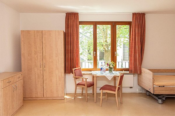 Geräumiges Zimmer mit Pflegebett,  einem Tisch vor einem Fenster, sowie Kommode und Kleiderschrank